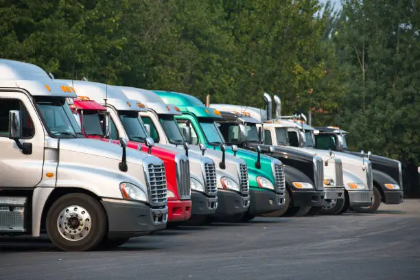 Photo of Different Big rig semi trucks tractors stand in row on parking lot with green trees