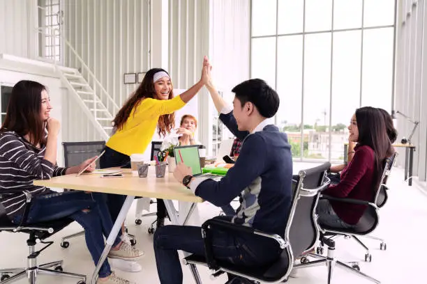 group of young multiethnic diverse people gesture hand high five, laughing and smiling together in brainstorm meeting at office. Casual business with startup teamwork community discussion concept.