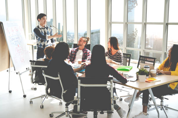 istock Young asian creative businessman standing and making presentation at modern office happy talking and brainstorming with teamwork on rear view and soft tone. Casual people business meeting concept. 1031251950
