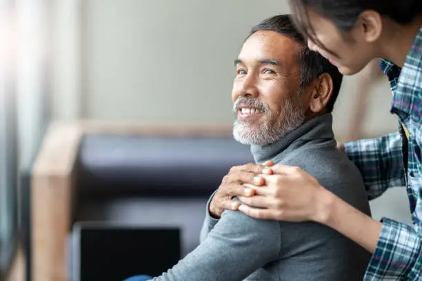 Photo of Smiling happy older asian father with stylish short beard touching daughter's hand on shoulder looking and talking together with love and care. Family relationship with bond and care concept.