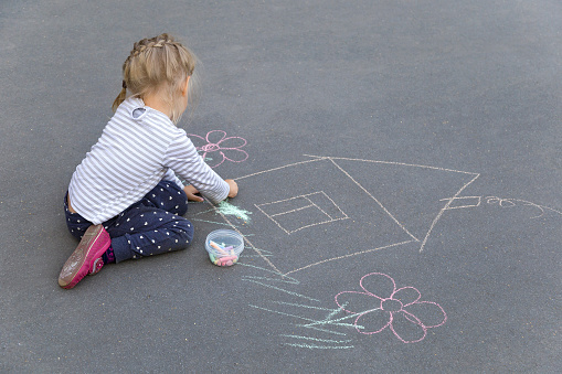 Little preschool caucasian girl drawing a house with chalks on the pavement