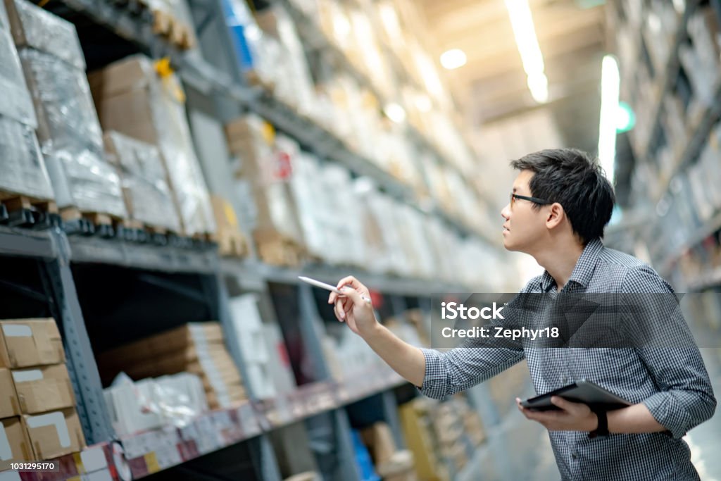 Young Asian man worker doing stocktaking of product in cardboard box on shelves in warehouse by using digital tablet and pen. Physical inventory count concept Warehouse Stock Photo