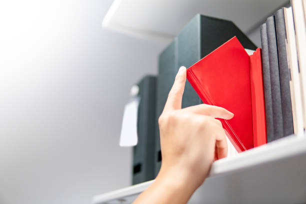 istock Male hand choosing and picking red book on white bookshelf in public library. Education research and self learning in university life concepts 1031295936