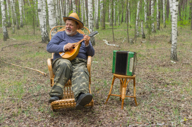 Senior singing and playing mandolin while sitting in a wicker rocking-chair in  birch forest