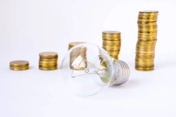 Light bulb is on background of stacks or columns of coins in ascending order. Concept photo saving electrical energy by people and industry, increase or decrease fees for the use of electricity