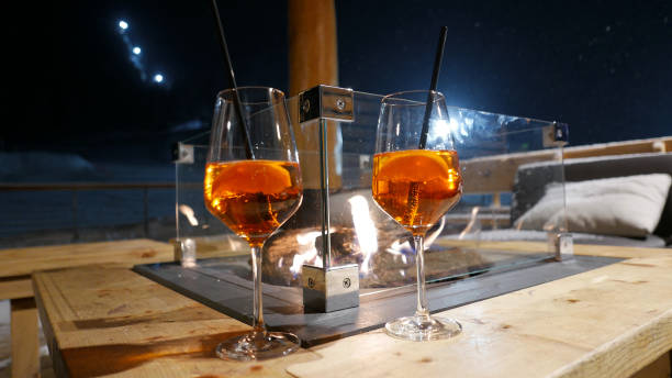 Two orange wine glasses with Spritz drinks and straws standing on winter terrace table with fire place in the middle