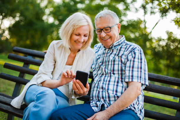 Senior man and woman in park Happy senior couple is using smartphone in park. senior adult women park bench 70s stock pictures, royalty-free photos & images