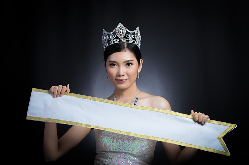 Portrait of Miss Pageant Beauty Contest in sequin Evening Ball Gown long dress sparkle light Diamond Crown, Asian Woman black hair style holding present Empty sash ribbon for country name copy space
