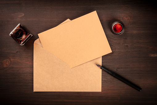 An overhead photo of an ink well with a brown craft card in an envelope, with an ink pen and copy space, on a dark wooden desk. A mockup for an invitation or greeting card