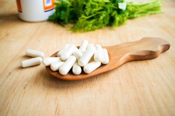 effets sur la santé de la vitamine allogène. capsules blanches de la vitamine k (phylloquinone) dans une cuillère en bois sur un fond d’une bouteille de comprimés et d’herbes vertes. suppléments de vitamines en cuillère sur la table en bois - letter k photos photos et images de collection