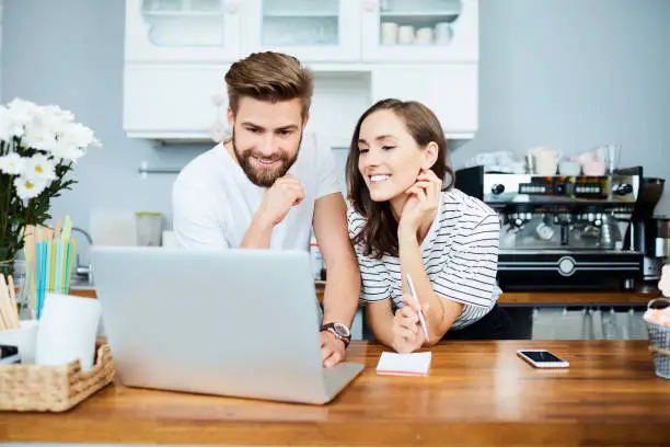 Couple of cheerful small business owners taking care of finances and payments using laptop and notepad in their restaurant