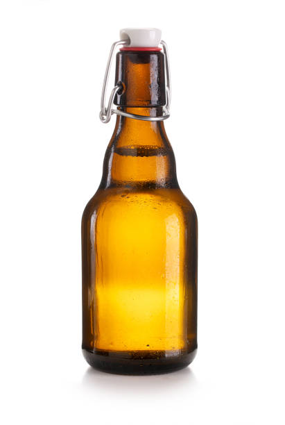 Full brown beer bottle isolated on white background with drops and condensation, back lit and showing a soft white reflection at right side. The bottle is opened and supporting a golden cap.