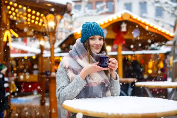 Beautiful young woman drinking hot punch, mulled wine on German Christmas market. Happy girl in winter clothes with lights on background on winter snow day in Berlin, Germany