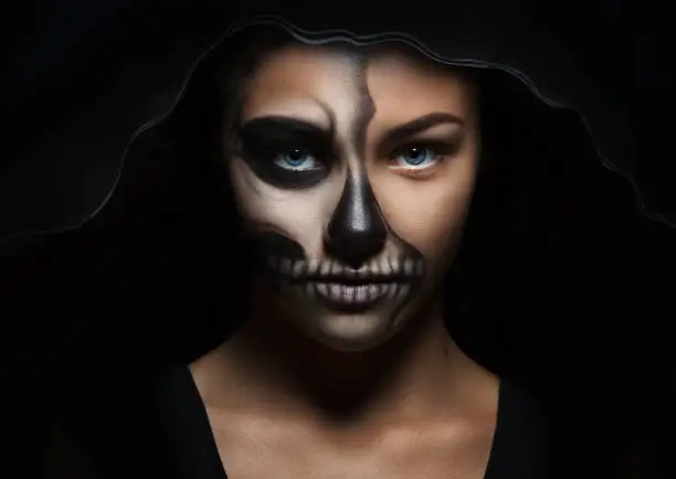 Photo of Halloween portrait of a young beautiful girl in a black hood. skeleton make-up half face