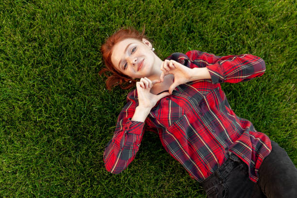 Beautiful red-haired young student with freckles lying on her back on the grass top view. Dressed in a red checked shirt, reveals a heart of fingers Love and positive.