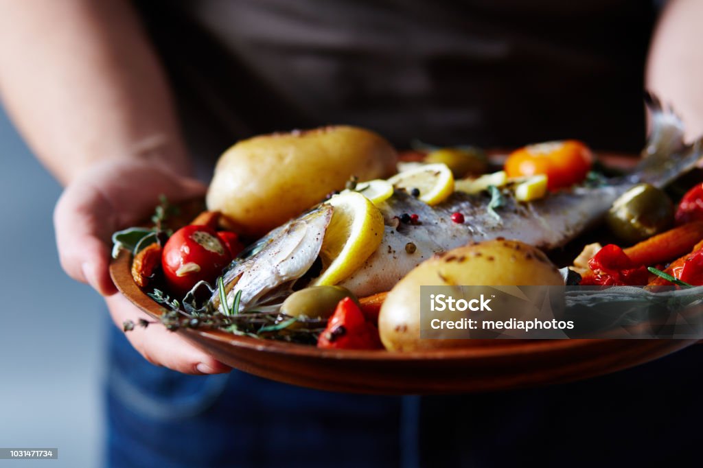 Appetizing Meal With Baked Fish Close-up view of unrecognizable man holding plate with delicious fish baked with potatoes, vegetables, spices and lemon Fish Stock Photo