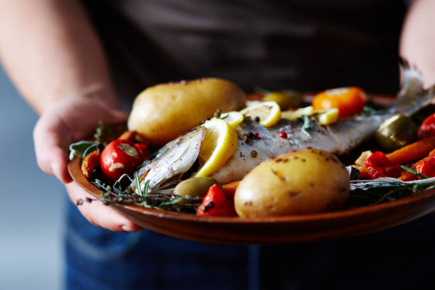 Appetizing Meal With Baked Fish Close-up view of unrecognizable man holding plate with delicious fish baked with potatoes, vegetables, spices and lemon roast dinner photos stock pictures, royalty-free photos & images