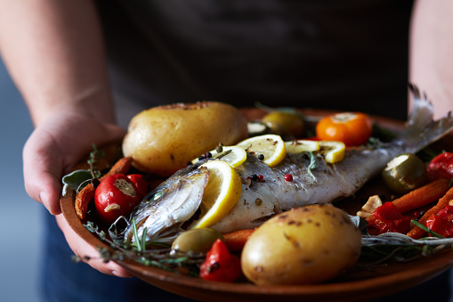 Close-up view of unrecognizable male hands holding tray with appetizing trout fish baked with potatoes, herbs and lemon