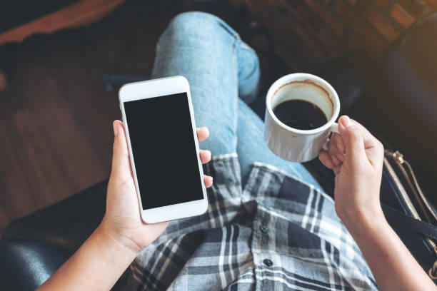 istock Top view mockup image of woman's hands holding white mobile phone with blank black desktop screen while drinking coffee in cafe 1031473762