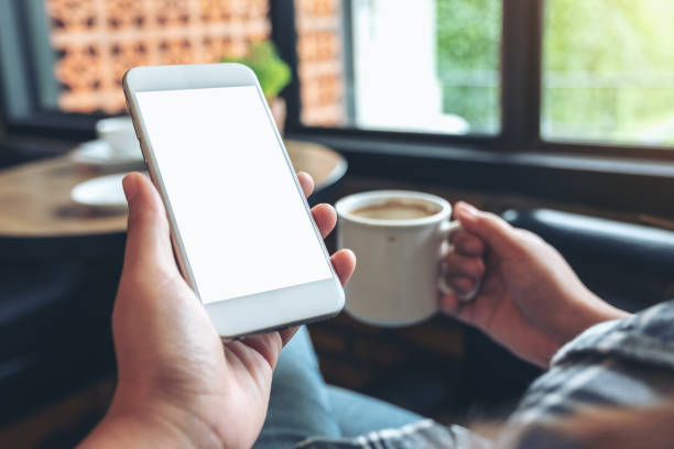 istock Mockup image of hands holding white mobile phone with blank screen while drinking coffee in modern cafe 1031476220