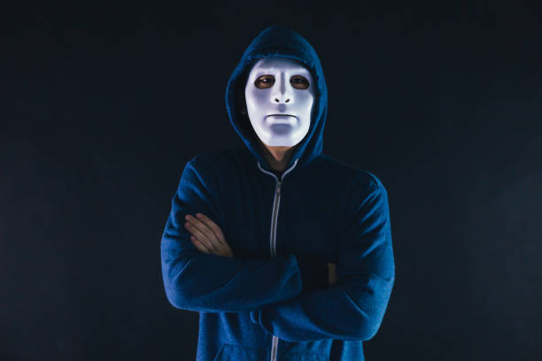 Anonymous masked man under hoodie with arms crossed isolated over dark background - incognito and mysterious criminal on internet activities concept. Anonymous masked man under hoodie with arms crossed isolated over dark background - incognito and mysterious criminal on internet activities concept creepy stalker stock pictures, royalty-free photos & images