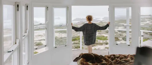 Woman standing near the balcony door of a beach house enjoying the sea view. Woman wearing a big sweater looking at the sea.