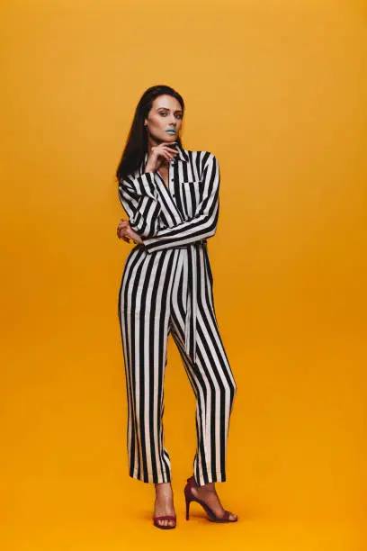 Full length portrait of female fashion model standing over orange background. Attractive woman with vivid make up in striped jumpsuit posing in studio.