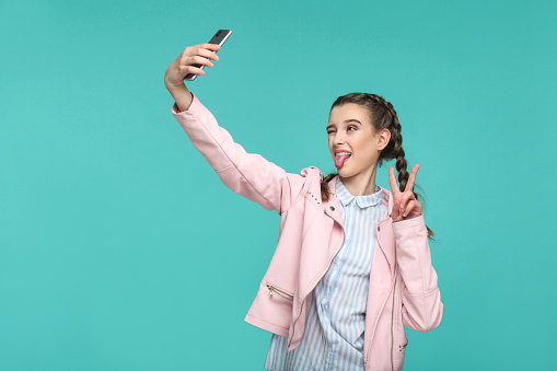 Profile side view of funny girl in casual style, pigtail hairs, standing, holding mobile smart phone and doing selfie with peace or victory sign and tongue out, studio shot on blue or green background