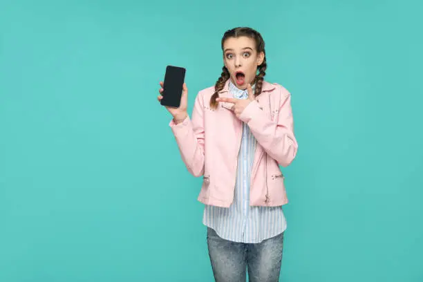 Photo of shocked funny girl in casual or hipster style, pigtail hairstyle, standing, holding and pointing at mobile display, screen with surprised face