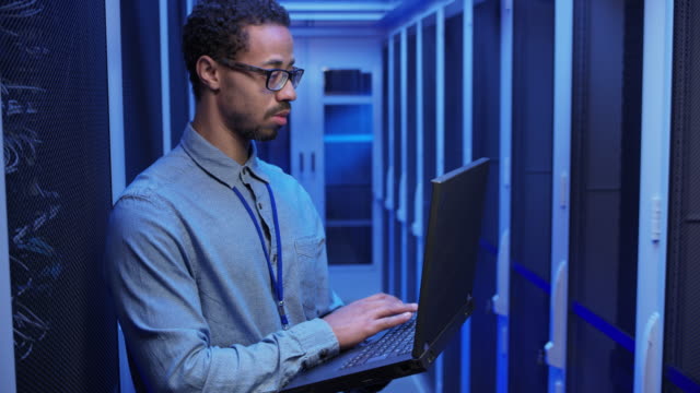 Male network engineer doing a system check standing in the server room with his laptop