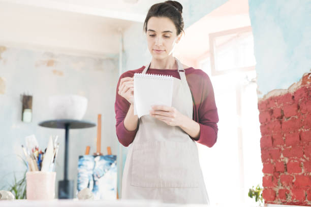 istock Serious pensive young female artist in apron drawing in notepad or checking notes while standing in workshop 1031584008