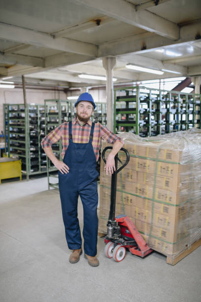 istock Content handsome young bearded mover leaning on pallet jack and looking at camera in factory warehouse 1031584454