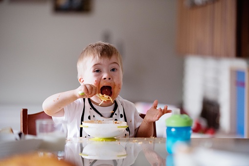Down's syndrome sweet boy eating spaghetti.  His face is dirty. He has blue eyes and blond hair. He is sitting at the table in the dining room. He is looking the camera. They wear pyjama short and t-shirts. The color and horizontal Photo was taken in Quebec Canada.