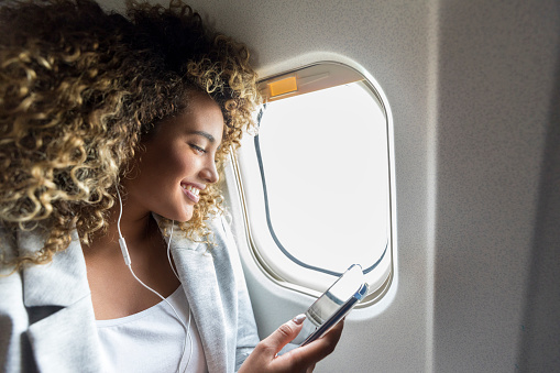 Confident woman selects music on smartphone to listen to during her flight. She is smiling cheerfully while wearing earbuds.
