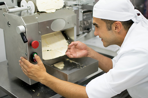 Latin american man at a pasta factory making lasagne sheets putting the dough through the press maker machine and looking very happy