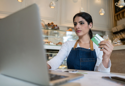 Serious female business owner of a bakery shopping online with her credit card using her laptop