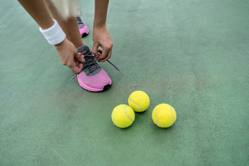 Female tennis player tying her shoelace at the tennis court standing next to the tennis balls