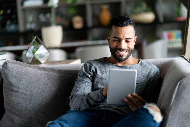 Cheerful latin american man relaxing on couch while looking at social media on tablet smiling Cheerful latin american man relaxing on couch while looking at social media on tablet smiling very happy e reader photos stock pictures, royalty-free photos & images
