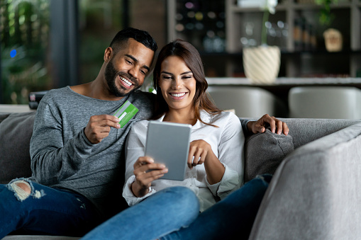 Beautiful adult couple at home relaxing on couch shopping online while man holds credit card both smiling very happy