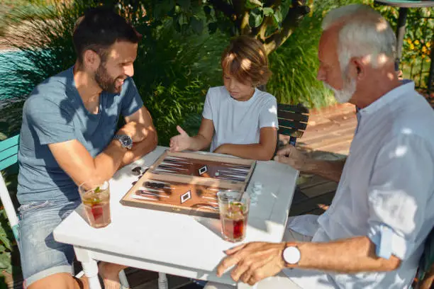 Three Generations Males Playing Backgammon Together on Summer Vacation. Sitting by the table, drinking refreshments and having fun.