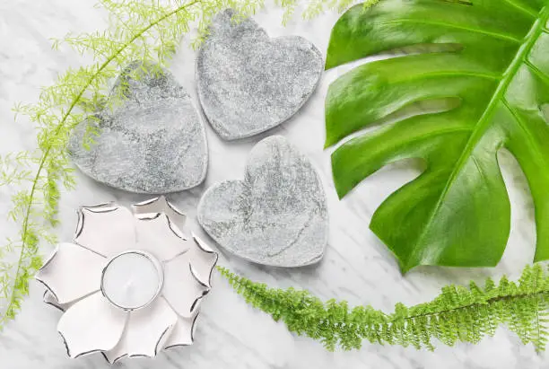 Green Monstera leaf, fern plants, stone hearts and lotus candle on marble background. Natural decor.