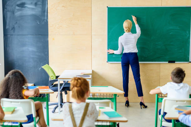 istock rear view of pupils looking at teacher while she writing on chalkboard 1031844564