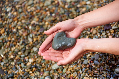 Wet female hands with a stone in the form of a heart against a background of wet pebbles on the beach.