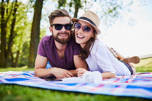 Young couple lying on blanket in park and looking at camera while wearing sunglasses