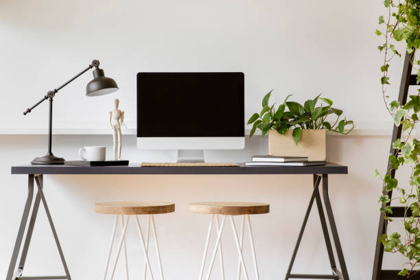 istock Wooden stools at desk with lamp, desktop computer and plant in white workspace interior. Real photo 1031886724