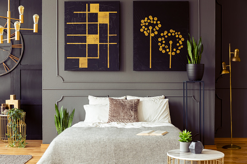 Real photo of a bedroom interior with big, black paintings with golden accents, double bed and plants
