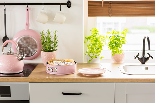 Pink kettle and cake on wooden countertop in bright kitchen interior with plants and pan. Real photo