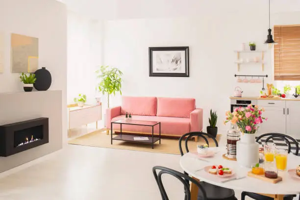 Flowers at table in white apartment interior with poster above pink settee near fireplace. Real photo