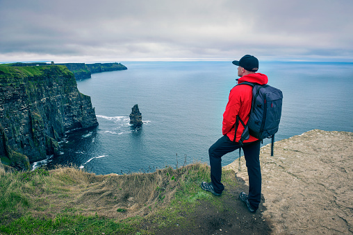 Young hiker with a backpack standing at the cliffs of Moher located at the edge of the Burren region in County Clare, Ireland.