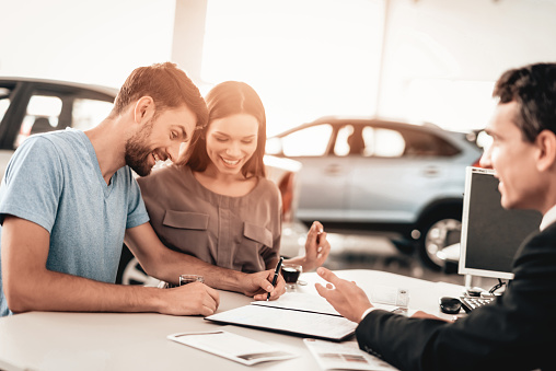 Happy Young Family Are Signing A Contract To Buy A Car. Dialogue With Dealer. Cheerful Customer. Automobile Salon. Make A Decision. Cup Of Coffee. End Of A Deal. Good Offer. Buyer And Seller.